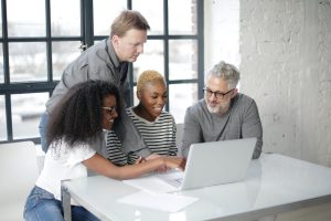 Three people sitting at a table looking at a laptop screen, one person standing and looking over