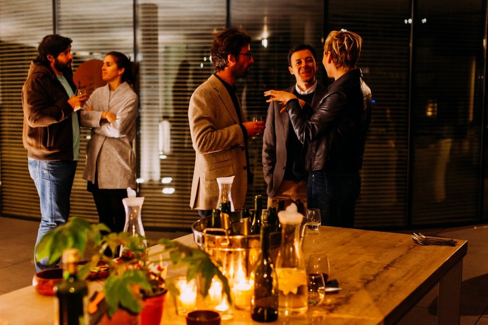 group of two men and woman talking and in background a woman talking to a man. Dim lighting in an office with drunks on the table in foreground