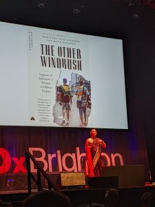 Dr María del Pilar Kaladeen on TedX Brighton stage with slide showing cover of book 'The Other Windrush'