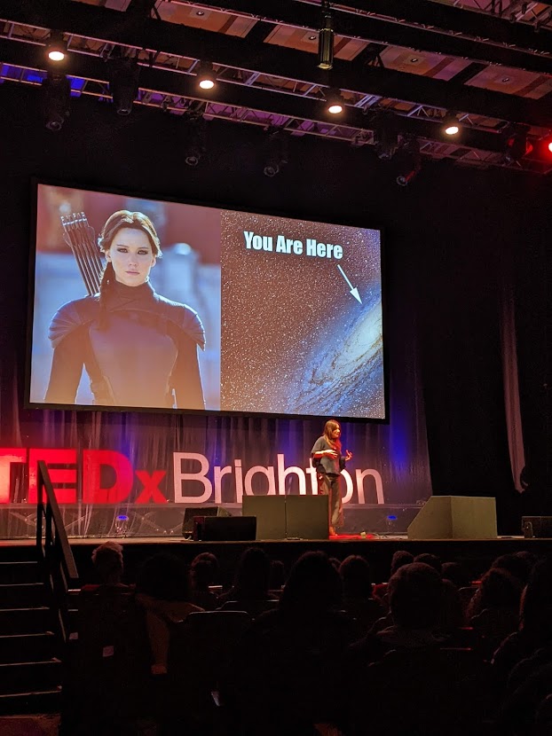 Dina Nayeri on TedX stage at Brighton Dome with picture of Katniss Everdeen and the universe behind her